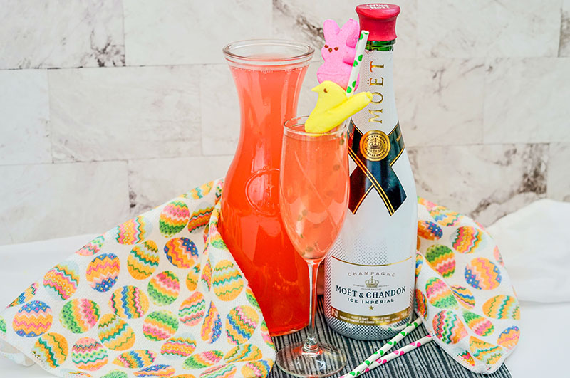 a photo of a peep mimosa next to a bottle of moet champagne and pink lemonade surrounded by a kitchen towel with easter eggs